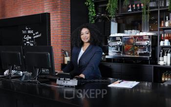 A professional at Teleportboulevard 110, Amsterdam Sloterdijk, stands behind a modern office setup with computers and coffee machines, ideal for office space rental.
