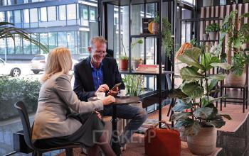 Two people engage in a discussion over coffee in a furnished office lobby at Teleportboulevard 110, Amsterdam Sloterdijk. This space offers a professional environment for office space rental.