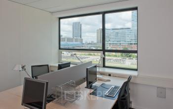 Bright office space rental in Amsterdam Sloterdijk with large window overlooking cityscape. Furnished with desks, chairs, and computer monitors, offering a modern workspace environment.