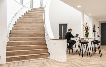 Modern office interior at Arlandaweg 92, Amsterdam Sloterdijk, featuring a stylish wooden staircase and a worker on a laptop at a high bar-style table with flowers.