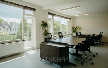 Bright office space at Overtoom 141, Amsterdam West, featuring large windows, modern desks, and office chairs, offering a pleasant workspace environment with natural light and greenery. Ideal for office space rental.