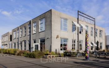 Exterior view of a modern office building at Rigakade 10, Amsterdam West, featuring large windows and a spacious outdoor area with benches. Suitable for businesses looking for office space rental in a vibrant location.