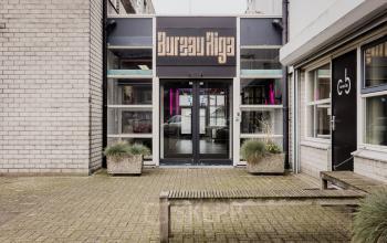 Exterior view of the office building at Rigakade 10, Amsterdam West, featuring a glass entrance with potted plants. Ideal for those seeking office space rental in a dynamic area.