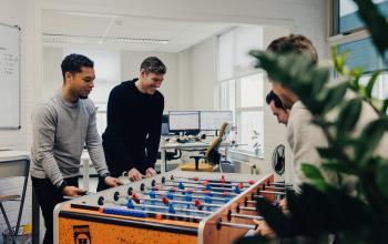 Office space at Rigakade 10, Amsterdam West, featuring a foosball table with colleagues engaged in a team activity near workstations. Ideal for office space rental seekers offering a vibrant atmosphere.