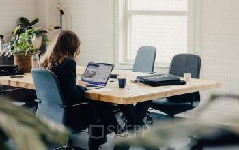Bright office space with large windows at Rigakade 10 in Amsterdam West, featuring a long wooden table and modern chairs, ideal for office space rental. A person works on a laptop, adding vibrancy to the space.