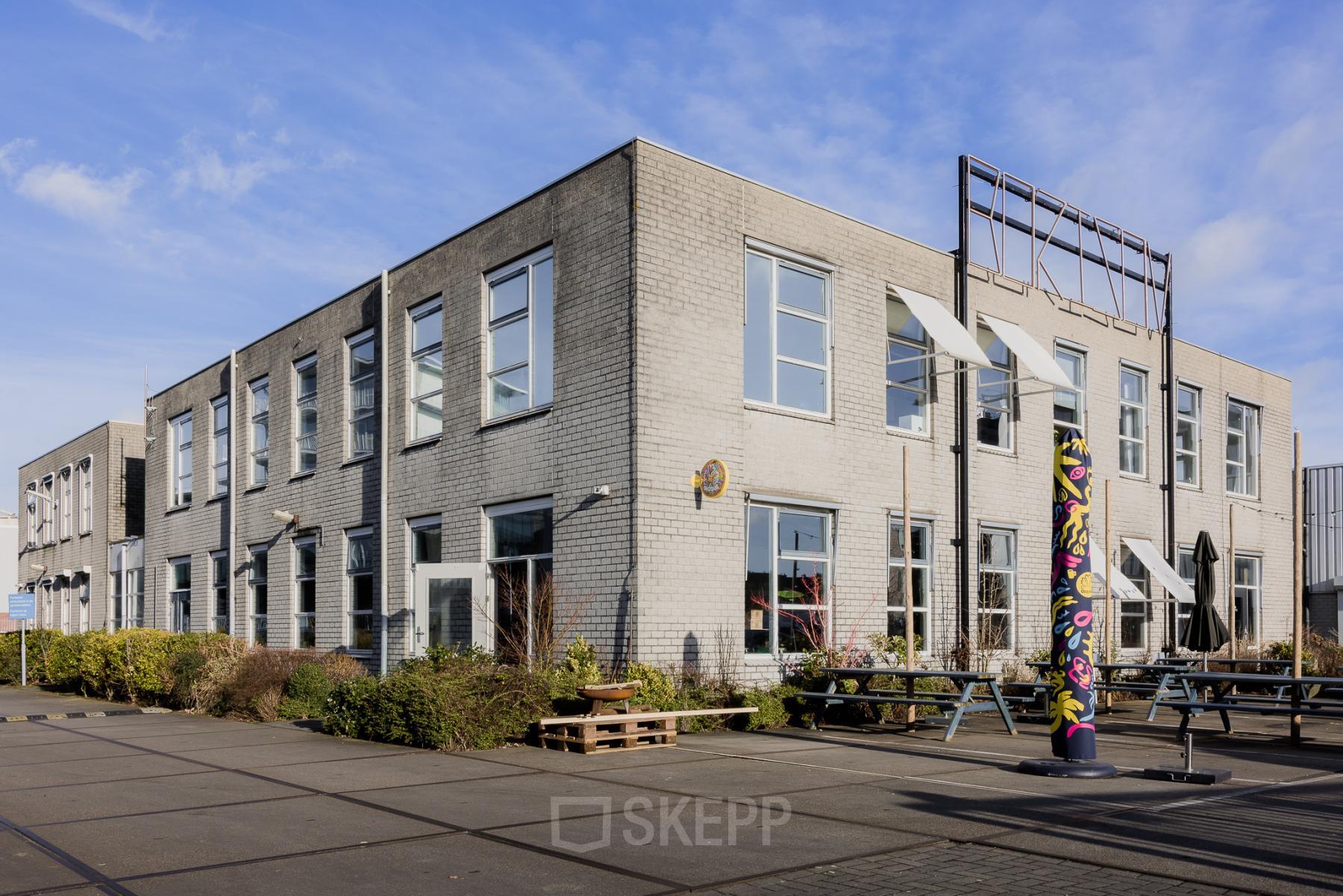 Exterior view of a modern office building at Rigakade 10, Amsterdam West, featuring large windows and a spacious outdoor area with benches. Suitable for businesses looking for office space rental in a vibrant location.