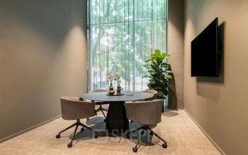 Furnished office space for rent at De Entree 201, Amsterdam South East, featuring a round table with chairs, a large window, and a plant.