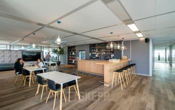 Modern office lounge in Amsterdam South East with sleek wood flooring, tables, and a central bar area, perfect for informal meetings. Ideal for those seeking office space rental.