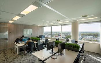 Bright workspace with large windows and modern furnishings at De Entree 230-266, Amsterdam South East; offering ample office space rental with city views. Two people are engaged in business activities at desks.