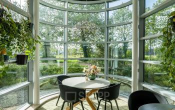 Bright circular meeting area in office at De Entree 230-266, Amsterdam South East, featuring floor-to-ceiling windows, lush plants, and modern furniture, ideal for office space rental.