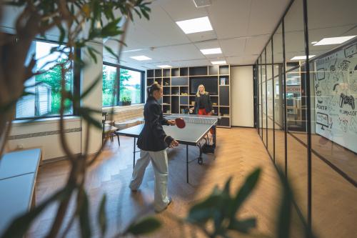 Two people playing table tennis in a well-lit office with wooden floors and large windows at Luttenbergweg 8, Amsterdam South East. Ideal for office space rental.