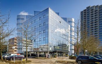 Modern glass office building located at Hogehilweg 8 in Amsterdam South East on a clear day, surrounded by parking spaces and trees.