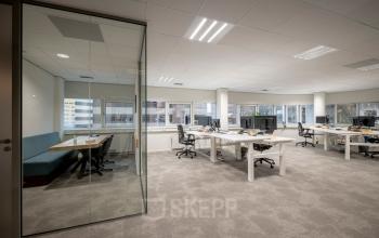 Modern office space rental at Hogehilweg 8, featuring open floor plan with workstations, glass-enclosed meeting area, and large windows for natural light.