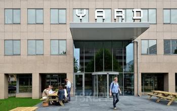 The exterior of the office building at Karspeldreef 8, Amsterdam South East, showcasing the main entrance with people engaged in a conversation near wooden benches in front of the building.
