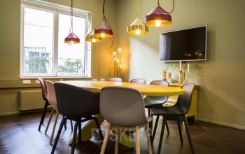 Contemporary meeting room with a wooden table, colorful chairs, pendant lighting, and a wall-mounted TV. Ideal for office space rental at Burgemeester Stramanweg 102 in Amsterdam South East.