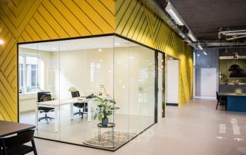 Modern office space rental featuring glass-walled workspaces and a sleek interior design at Burgemeester Stramanweg 102, Amsterdam South East. The space includes multiple desks, chairs, and a plant for a lively atmosphere.