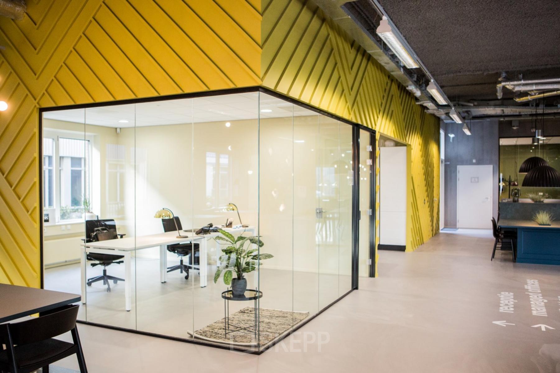 Modern office space rental featuring glass-walled workspaces and a sleek interior design at Burgemeester Stramanweg 102, Amsterdam South East. The space includes multiple desks, chairs, and a plant for a lively atmosphere.