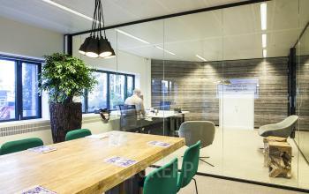 Office space at De Entree 99-197, Amsterdam South East with a modern conference table, green chairs, glass walls, and a person working at a desk.