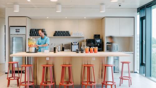 Modern pantry area at an office located at Strawinskylaan 4117, Amsterdam Zuidas, with stools, countertops, and a worker preparing beverages.