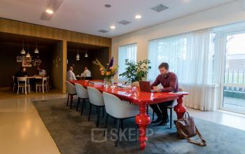 Modern office space in Amsterdam Zuidas with a vibrant red table, comfortable chairs, and large windows offering natural light. Professionals are engaged in work at their laptops. Ideal office space rental.