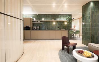 Modern office space available for rent in Amsterdam Zuidas, featuring a furnished lounge area with seating and a kitchenette. A person is engaged in a business activity at a table.