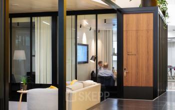 Modern office space at Fred. Roeskestraat 115 in Amsterdam Zuidas, featuring glass walls, wooden door, and comfortable seating, ideal for office space rental with meeting area and digital presentation screen.
