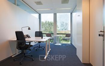 Modern office space rental at Gustav Mahlerplein 2, Amsterdam Zuidas, featuring large windows, work desks, office chairs, and a small meeting area with a view of greenery.
