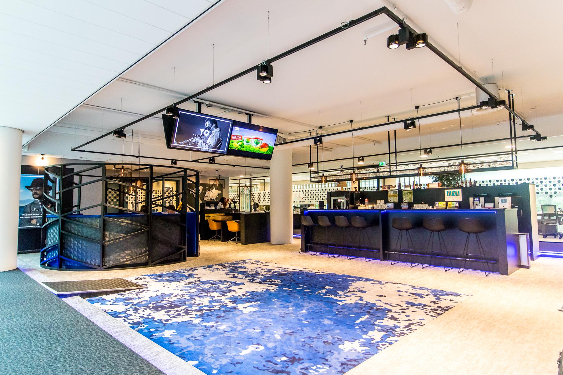 Bright, furnished office space available for rent at Gustav Mahlerplein in Amsterdam Zuidas, featuring a stylish open floorplan with a vibrant blue carpet and modern seating area.