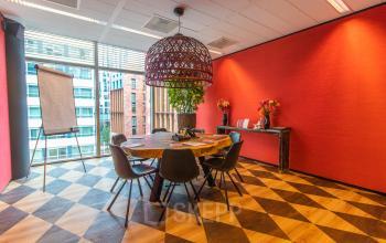 Bright conference room in Amsterdam Zuidas, featuring a large round table with chairs, vibrant red walls, and a large window overlooking a cityscape. Ideal for office space rental.