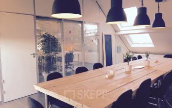 Bright office interior at Grote Hondstraat 44, Antwerp Zurenborg with large wooden table, plants, pendant lights, and adjacent workspace visible through glass partition.