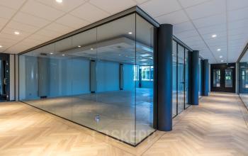 Spacious office space available for rent at Landdrostlaan 51 in Apeldoorn, featuring a modern glass-walled interior with ample natural light and sleek flooring.