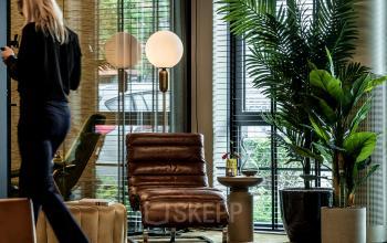Modern furnished office space in Apeldoorn with stylish seating and plants. A person is walking while using a smartphone, suggesting business activity.