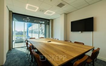 Modern conference room available for office space rental at Prins Willem Alexanderlaan 701, Apeldoorn, featuring a large wooden table, chairs, a wall-mounted screen, and a bright window view.