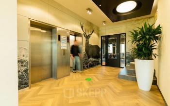 Modern office entrance at Prins Willem Alexanderlaan 701, Apeldoorn, featuring elegant wood flooring, stylish artwork, and a person exiting the elevator. Ideal for office space rental.