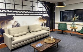 A modern office lounge in Apeldoorn, featuring comfortable seating and stylish decor, ideal for office space rental. The room has a sofa, chairs, a coffee table, and decorative wall art.