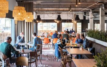 Lively office space at Oude Apeldoornseweg 41-45, Apeldoorn, featuring people engaged in discussions and dining at wooden tables with vibrant chairs.