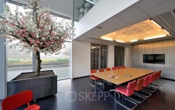 Modern conference room at Laan van Malkenschoten 40, Apeldoorn with large table, red chairs, and a decorative indoor tree.