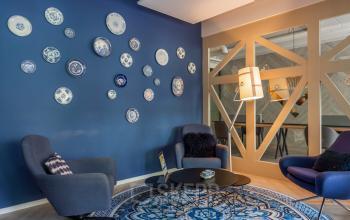 Cozy furnished lounge area with blue wall, decorative plates, blue chairs, round table, and modern lamp in an office space rental at Roggestraat 111, Apeldoorn.