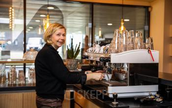 Person operating a coffee machine in the pantry area of the office at Roggestraat 111, Apeldoorn. Modern lighting and glassware are visible.