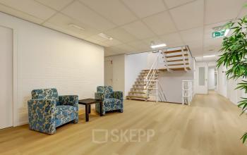 Interior view of Bruningweg 23, Arnhem IJsseloord, featuring a minimalist lounge area with two patterned chairs, a wooden table, and a staircase leading to office space rental opportunities.