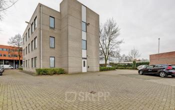 Exterior view of an office building at Bruningweg 23 in Arnhem IJsseloord, featuring a brick facade with multiple windows and a parking area. Ideal for businesses looking to rent office space.