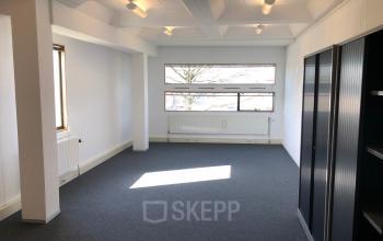 Bright office space with blue carpet, large windows, overhead lighting, and shelving units on Simon Stevinweg 27, Arnhem Oost, ideal for office space rental.