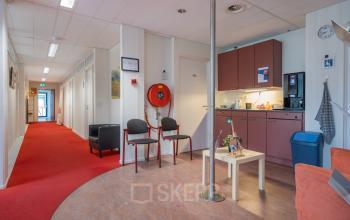 Interior view of a cozy office space at Overcingellaan 17, Assen, featuring a small kitchenette and lounge area with red carpet and a couple of chairs, ideal for office space rental.
