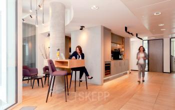 Moderner Büroraum im Upper West Tower, Kurfürstendamm 11, Berlin Charlottenburg. Eine Person arbeitet am Tisch, während eine andere den Raum durchquert. Ein einladender Bereich zum Büro mieten.