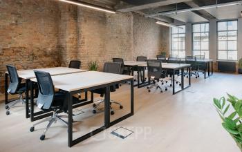 Büroraum mieten im Bürogebäude an der Köpenicker Straße 40-41, Berlin Friedrichshain. Geräumiger Arbeitsbereich mit mehreren Schreibtischen und Stühlen, freiliegende Backsteinwand und große Fenster sorgen für natürliche Beleuchtung.