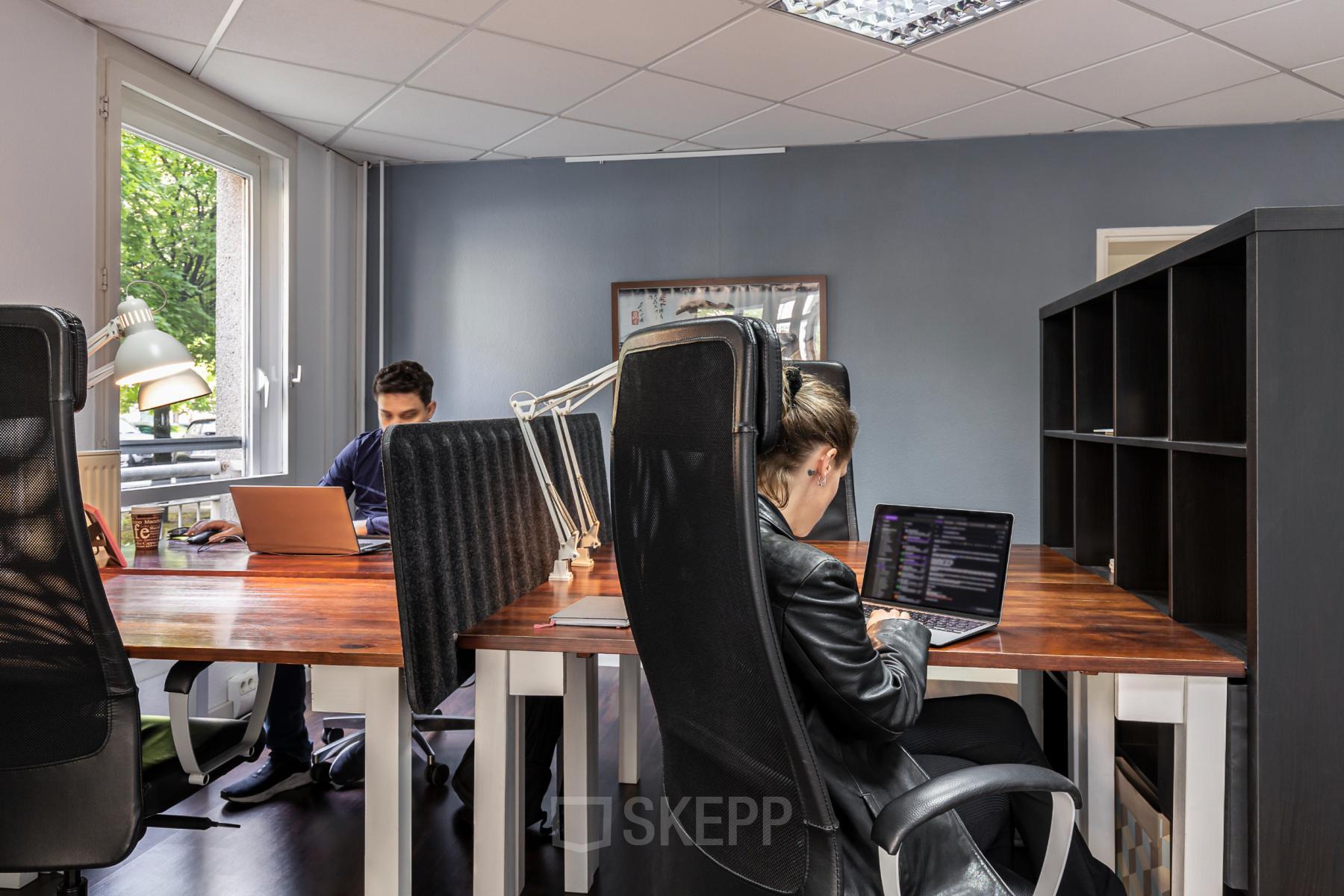 Cozy office space rental in Berlin Mitte featuring wooden desks, modern chairs, and large windows offering natural light. Two individuals are engaged in work at their laptops, fostering a productive atmosphere.