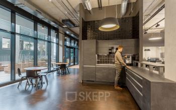 Modern office kitchen at Hussitenstraße 32, Berlin Mitte, with a person preparing coffee by the countertop, large windows providing natural light, and sleek furnishings, ideal for office space rental.