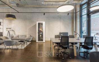 Modern office space for rent at Hussitenstraße 32, Berlin Mitte, featuring large windows, open workstations, and a cozy lounge area. A person is seen making a phone call in a booth.