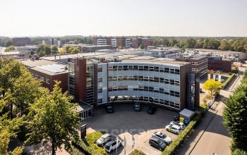 Exterior view of office space rental at De Maas 8, Best, showcasing a modern, multi-floor building surrounded by greenery and a parking lot.