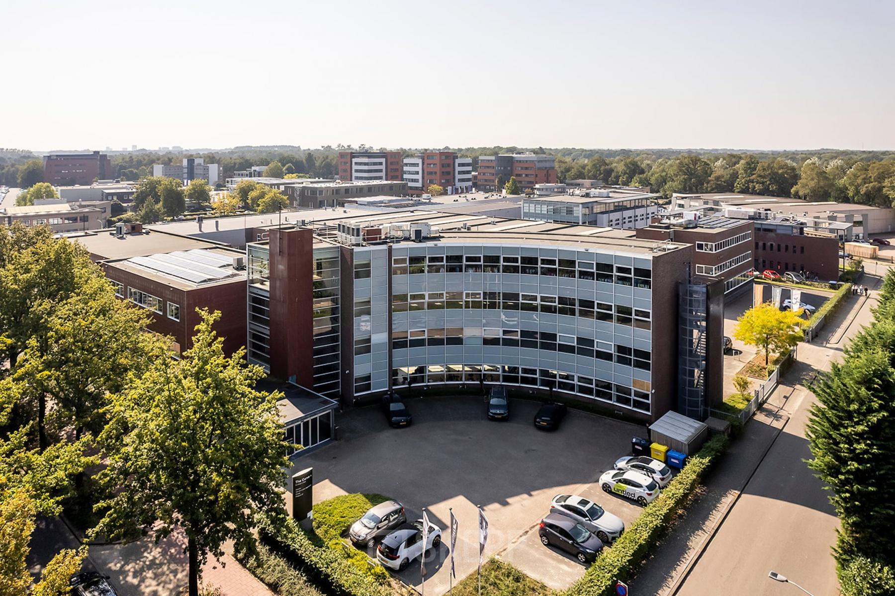 Exterior view of office space rental at De Maas 8, Best, showcasing a modern, multi-floor building surrounded by greenery and a parking lot.